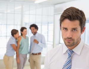 Colleagues gossiping with sad young businessman in foreground at a bright office